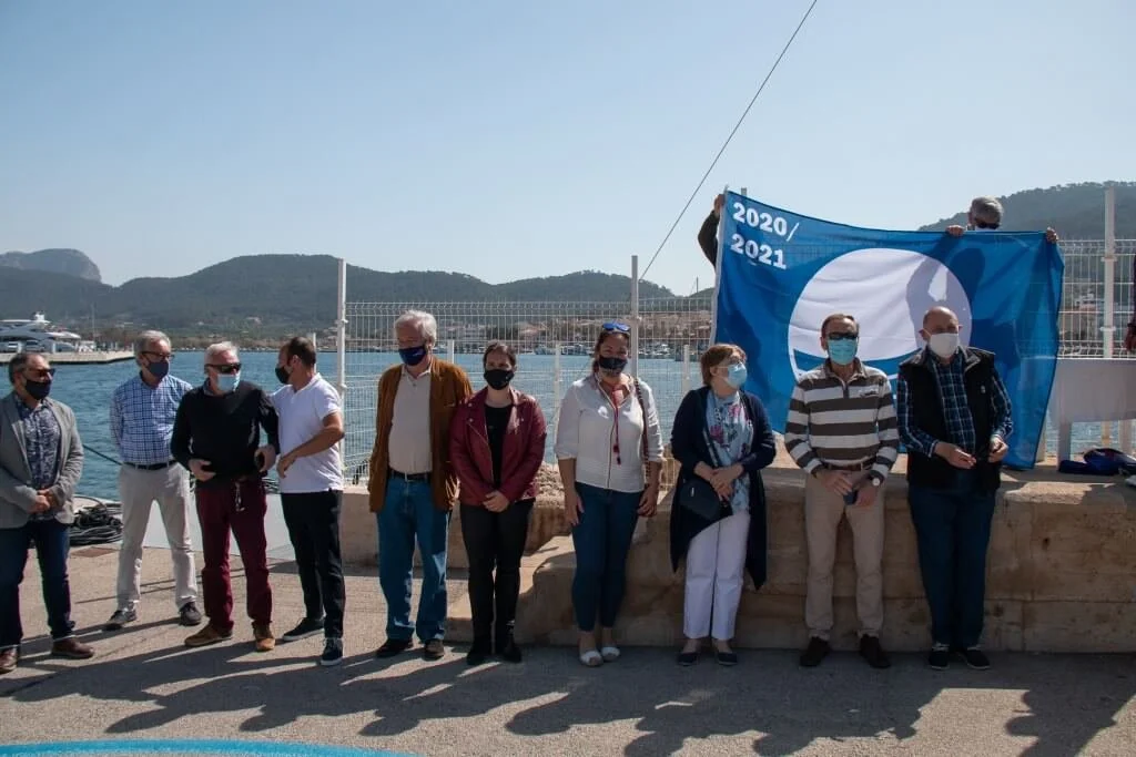 Grupo de personas de pie junto a un muro con bandera azul que dice 2020/2021, en un puerto con montañas de fondo, algunos con mascarillas.