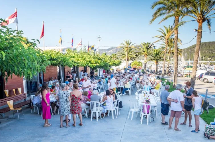 Personas disfrutando una comida al aire libre en una terraza con palmeras y banderas. 