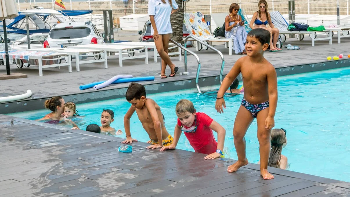 Niños y niñas en una piscina jugando y nadando, algunos están en el borde y otros en el agua, mientras unas personas en el fondo están sentadas y usando teléfonos