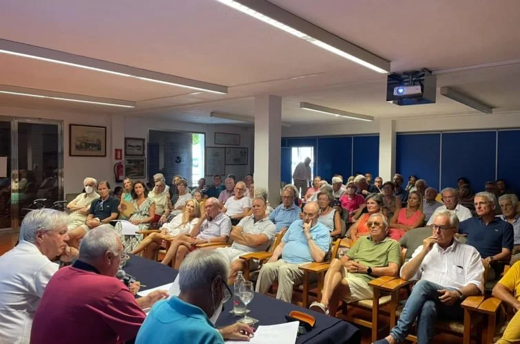 Reunión de personas en una sala, todas sentadas y prestando atención a una mesa frente a ellos donde hay varias personas y documentos.