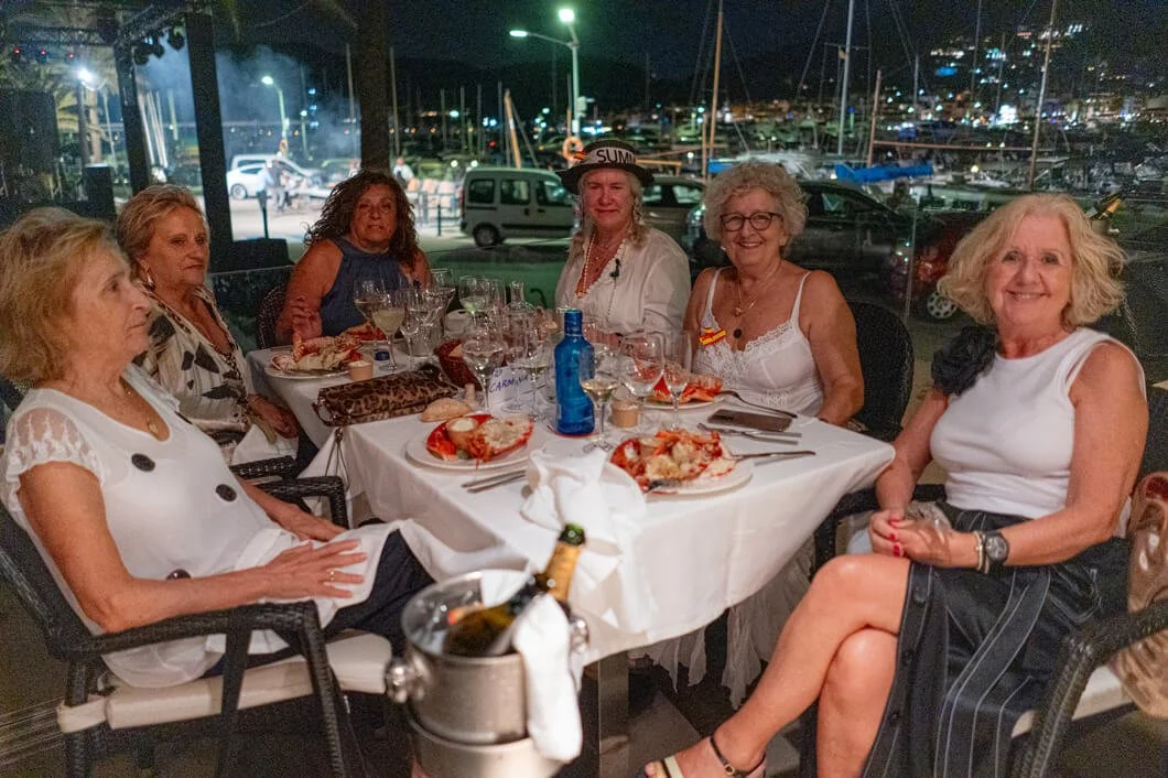 Grupo de mujeres mayores en una cena en un restaurante con vista al puerto y barcos al fondo de noche.