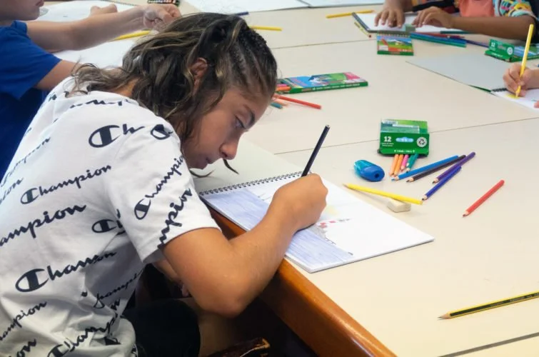 Una niña en un aula escribiendo en su cuaderno con varios útiles escolares dispersos en la mesa.