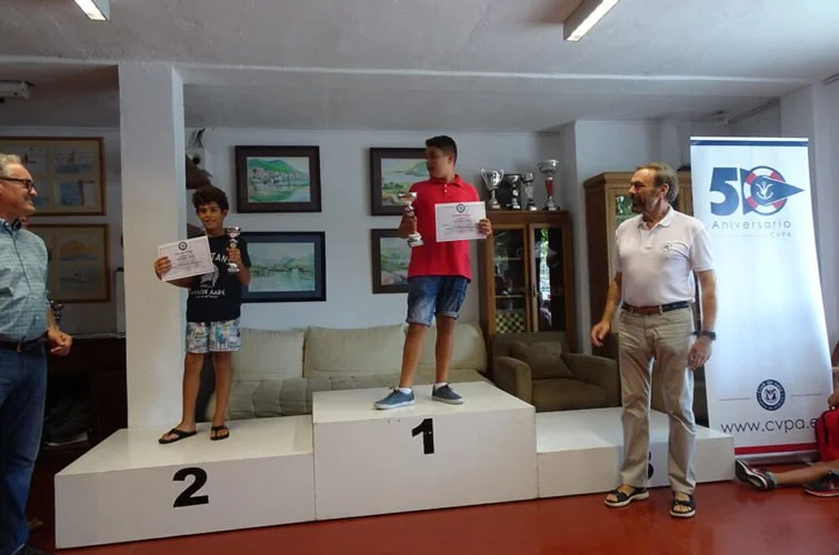 Niños en un podio de premiación, con el niño en el primer lugar sosteniendo un trofeo, y otros niños y adultos presentes en la ceremonia