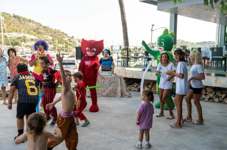 Niños y adultos en una fiesta al aire libre con payasos y personajes de dibujos animados, bailando y jugando.