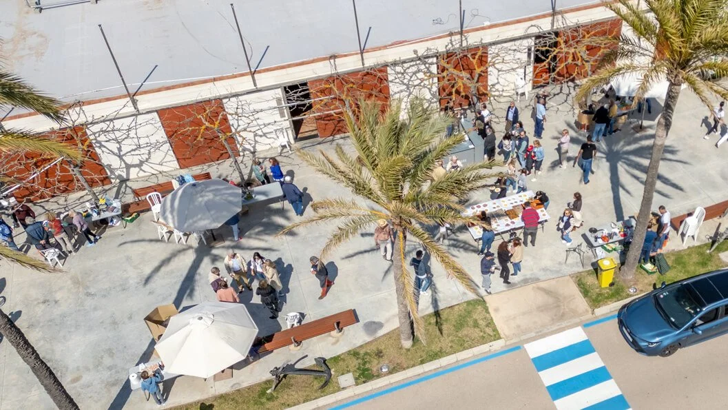 Vista aérea de una calle concurrida con personas en una plaza, palmeras, mesas con comida y un puesto con sombrilla, en un día soleado.