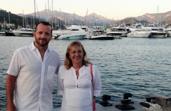Una pareja sonriente de pie junto a un puerto con botes y yates, con montañas al fondo al atardecer.