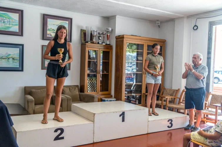 Dos jóvenes en el podio recibiendo trofeos, acompañados por un hombre que los felicita en una ceremonia en interior.