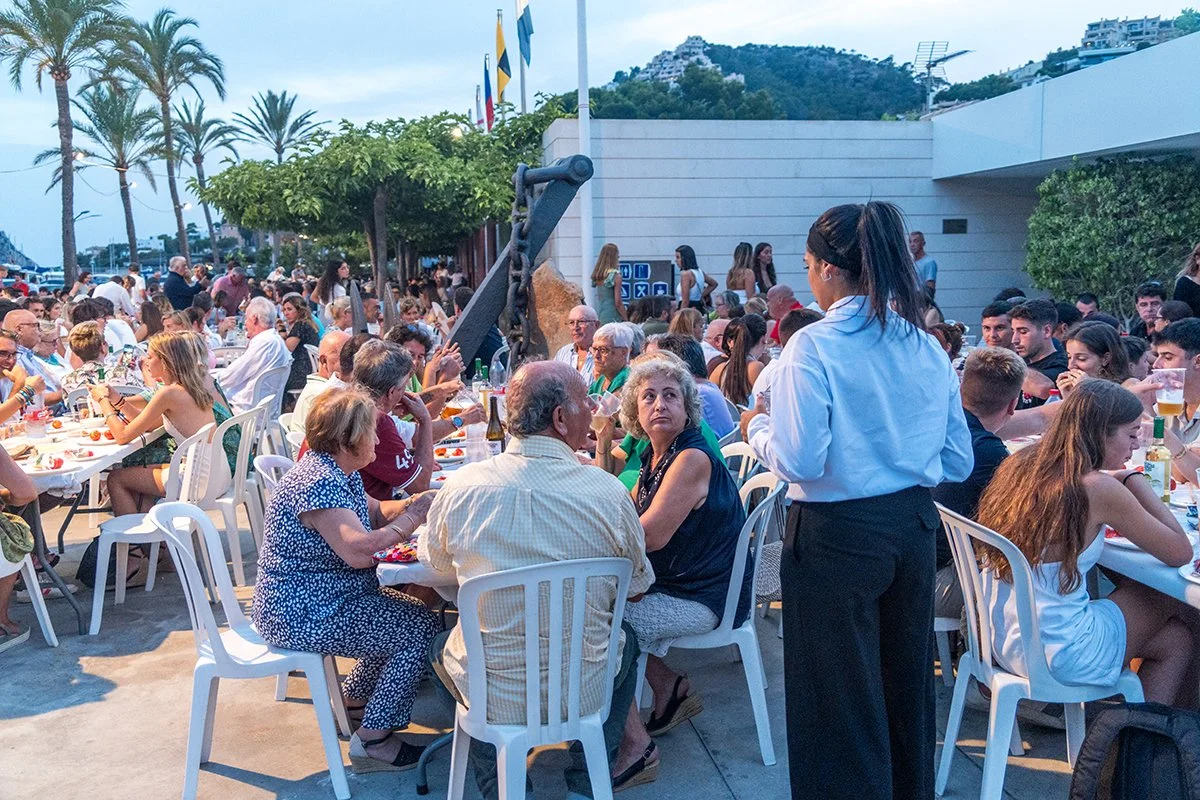 Reunión social al aire libre con muchas personas sentadas en mesas comiendo y bebiendo, con una mujer de pie atendiendo a los invitados, árbol y montañas al fondo.