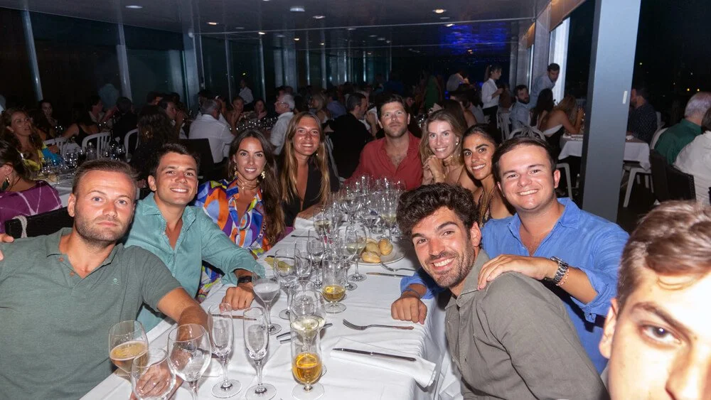 Grupo de personas disfrutando de una comida en un restaurante elegante durante la noche, con mucha gente en el fondo.
