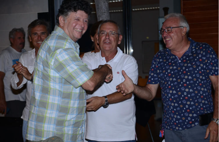 Tres hombres sonriendo y dándose la mano en un ambiente interior, parece una reunión social o celebración.