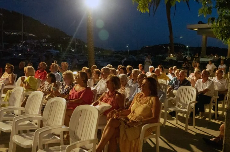 Personas sentadas en sillas blancas al aire libre durante la noche, algunas conversando y otras escuchando, en un evento en un puerto o mar, con árboles y montañas al fondo.