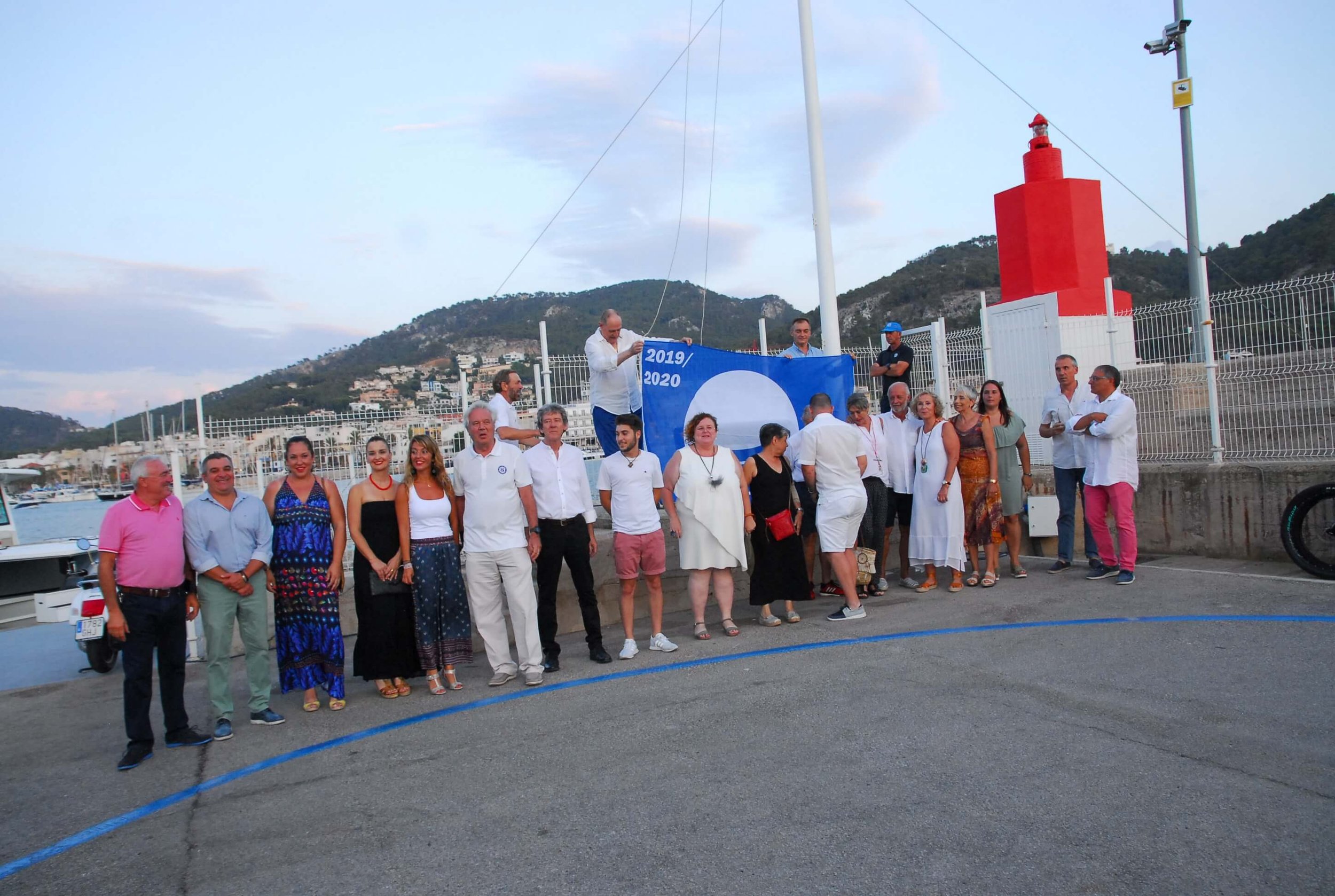 Grupo de personas en una reunión al aire libre en el puerto, algunos sosteniendo una bandera con los años 2019/2020, con montañas y un faro rojo al fondo.