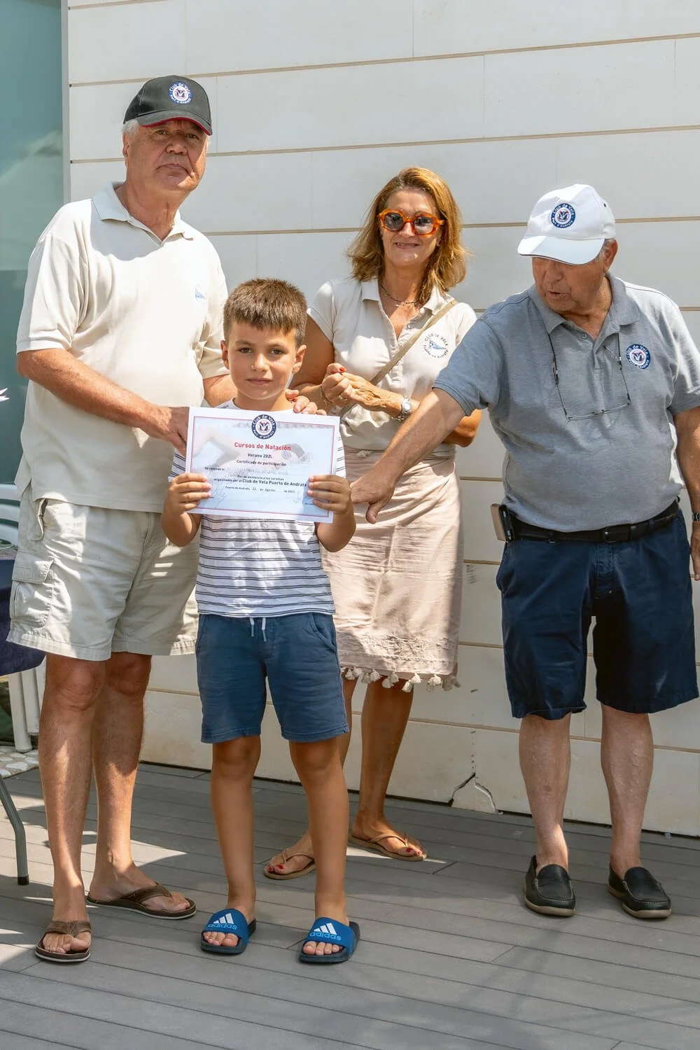 Un niño sostiene un certificado de participación en un curso de natación, rodeado de adultos que parecen ser instructores o coordinadores del evento. Todos están al aire libre, frente a una pared de color claro.