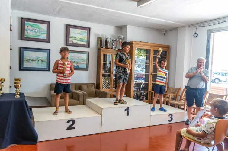 Niños en un podio de premios en una ceremonia de entrega, con un niño en primer lugar y otros en segundo y tercer lugar, mientras un adulto observa y una niña en el público lee un papel.
