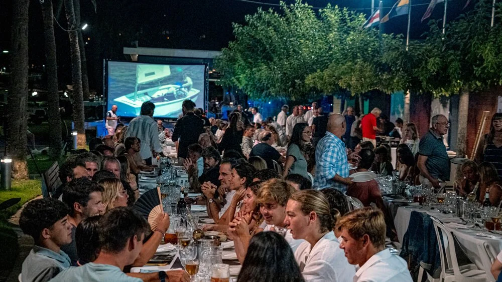 Cena al aire libre con muchas personas comiendo y bebiendo, una pantalla gigante muestra una concertina y en el fondo hay árboles y banderas.