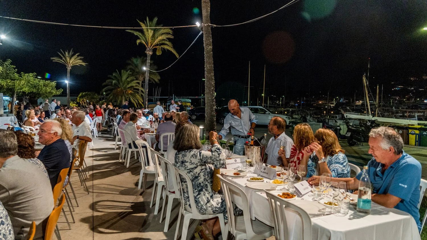 Cena de una cena al aire libre en la noche junto a un muelle con barcos. Personas sentadas en mesas con comida y copas, algunos siendo atendidos por camareros. Palmas y luces en el fondo.