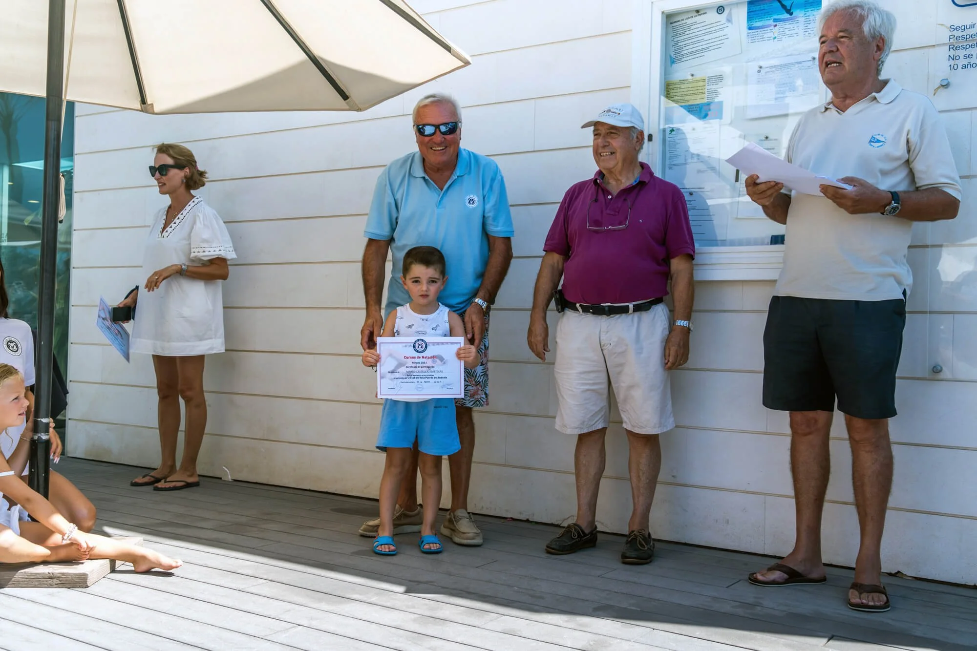 Un grupo de personas en una ceremonia de premiación en una piscina cubierta, con un niño sosteniendo un diploma y adultos sonriendo junto a una pared con avisos y cartelera informativa.