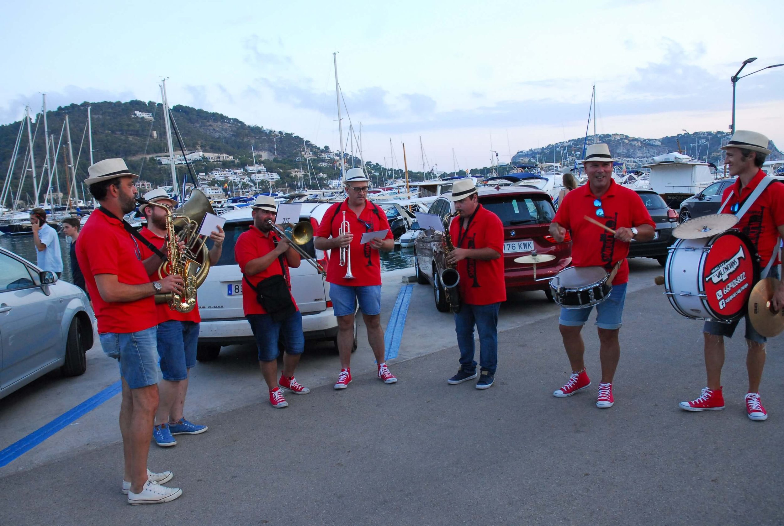 Grupo de músicos tocando en un muelle con yates y colinas en el fondo, todos llevan camisetas rojas y sombreros blancos, algunos con gafas y calzado deportivo rojo o azul.