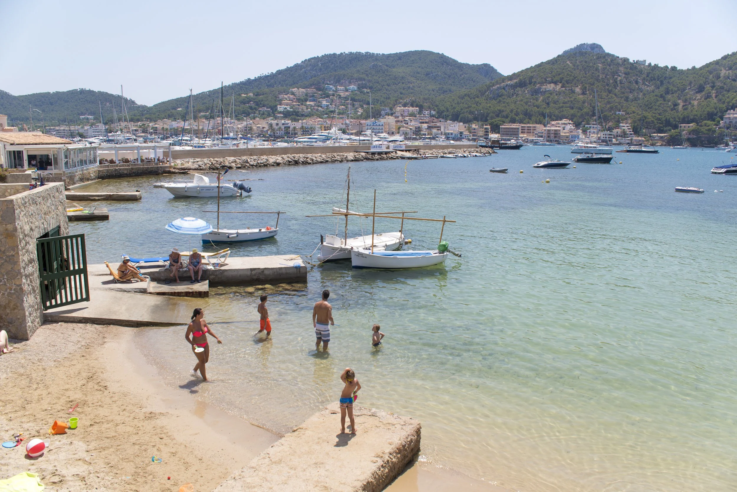 Vista de un puerto con barcos en el agua y una ciudad en las colinas al fondo, con personas disfrutando en la playa en primer plano.