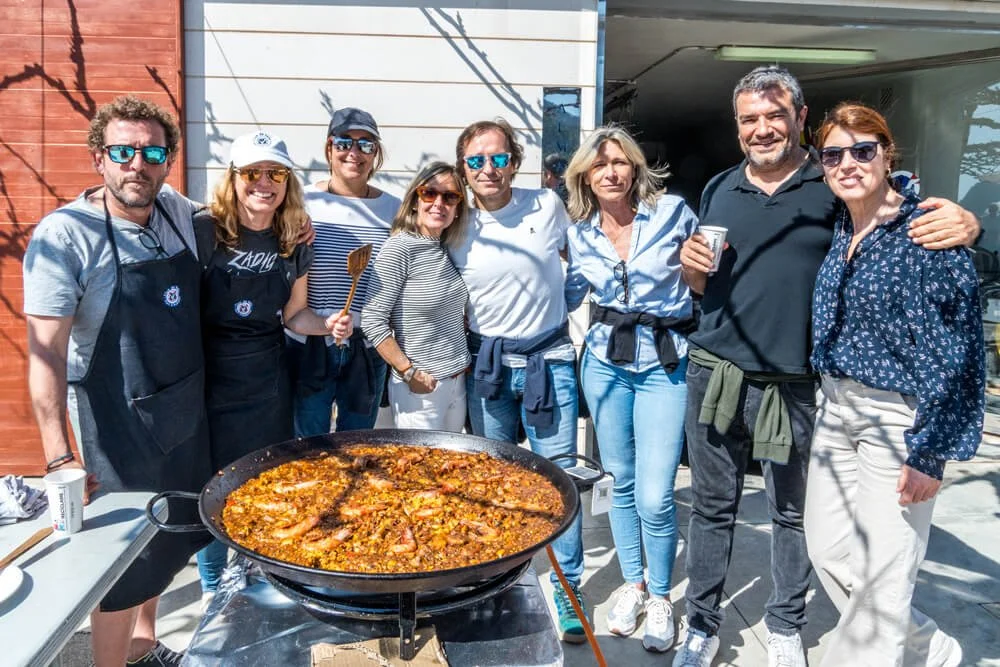 Grupo de ocho personas sonriendo, con una paella gigante frente a ellos, en un espacio abierto con fondo de pared de madera y una estructura de garaje.