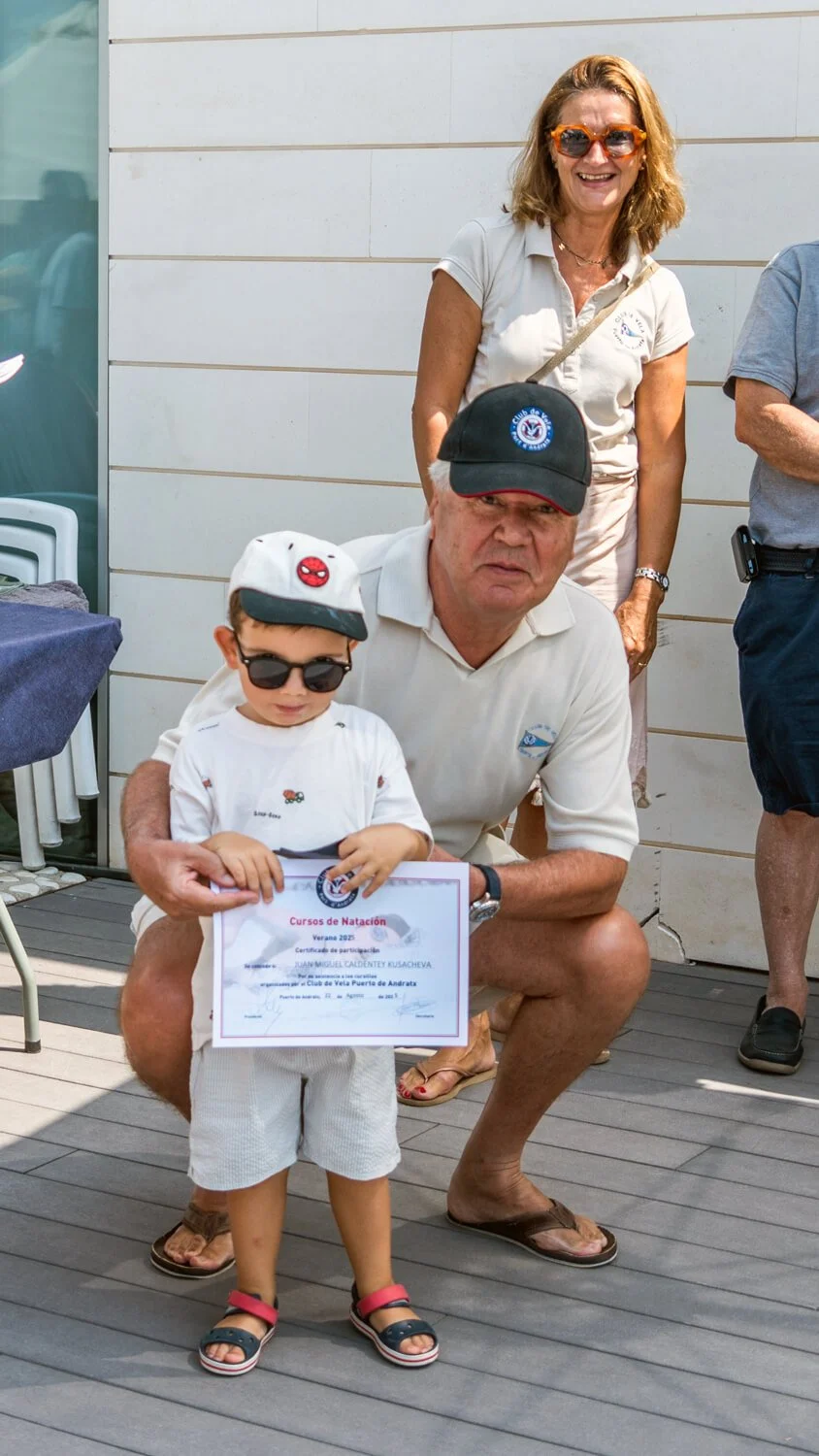 Un niño con gafas y gorra sosteniendo un certificado de participación en cursos de natación, acompañado por un hombre mayor que está junto a él, en un evento al aire libre.