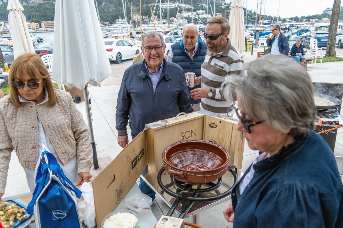 Grupo de personas en un evento al aire libre en un muelle con barcos y autos cerca, cocinando y comiendo en un puesto de comida, con una olla grande de ingredientes en el centro.