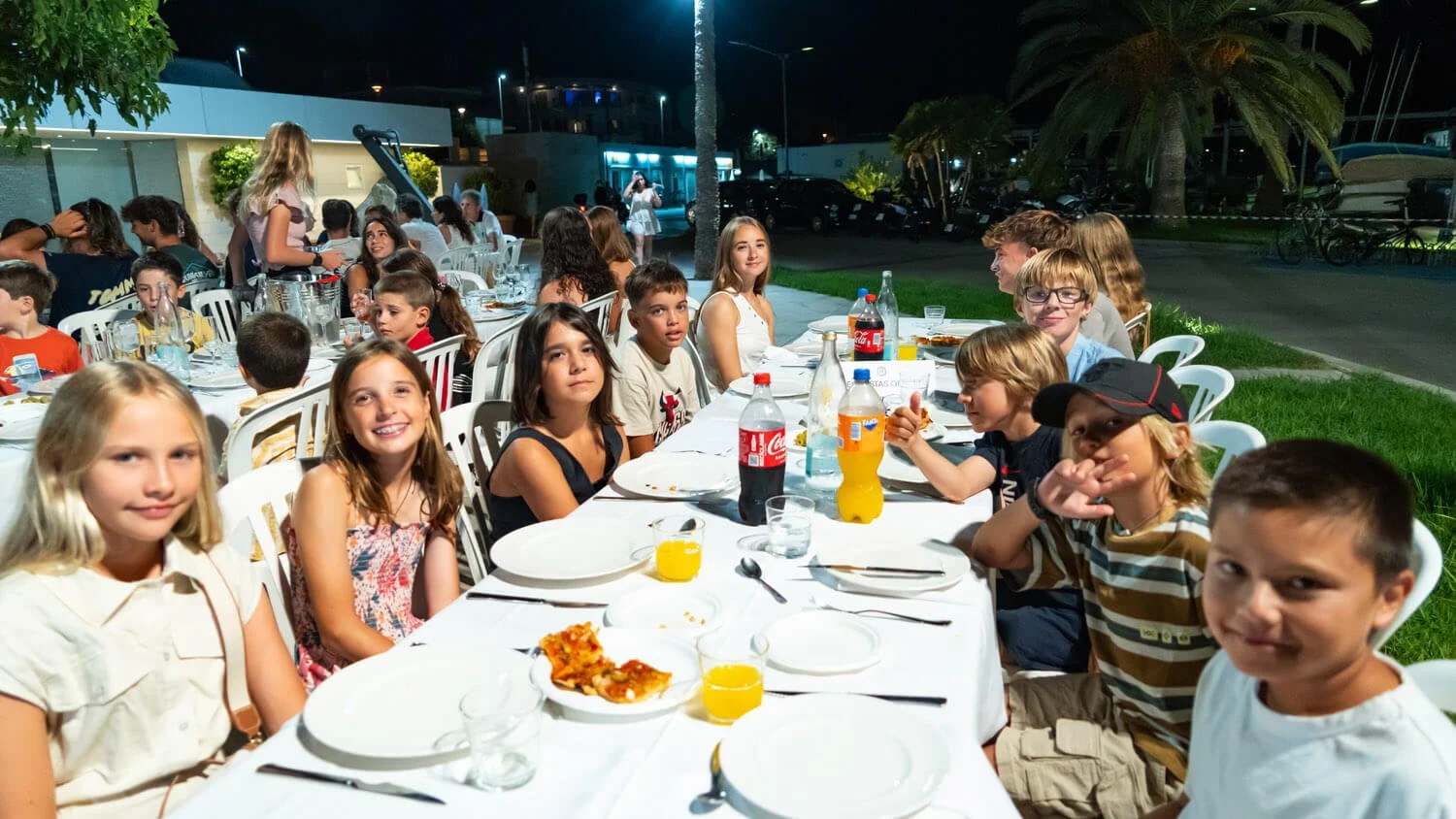 Grupo de niños y niñas sentados en una mesa durante una fiesta al aire libre en la noche, celebrando con comida y bebidas.