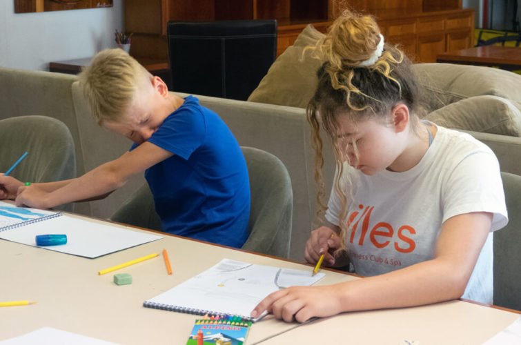 Dos niños sentados en una mesa, dibujando con crayones y lápices, aparentemente cansados o aburridos.