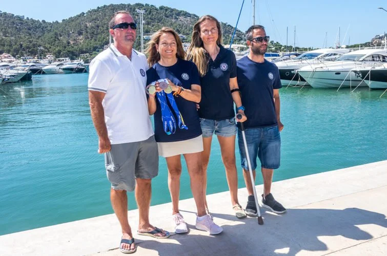 Cuatro personas de pie junto al muelle con botes y yates en el fondo, en un día soleado.