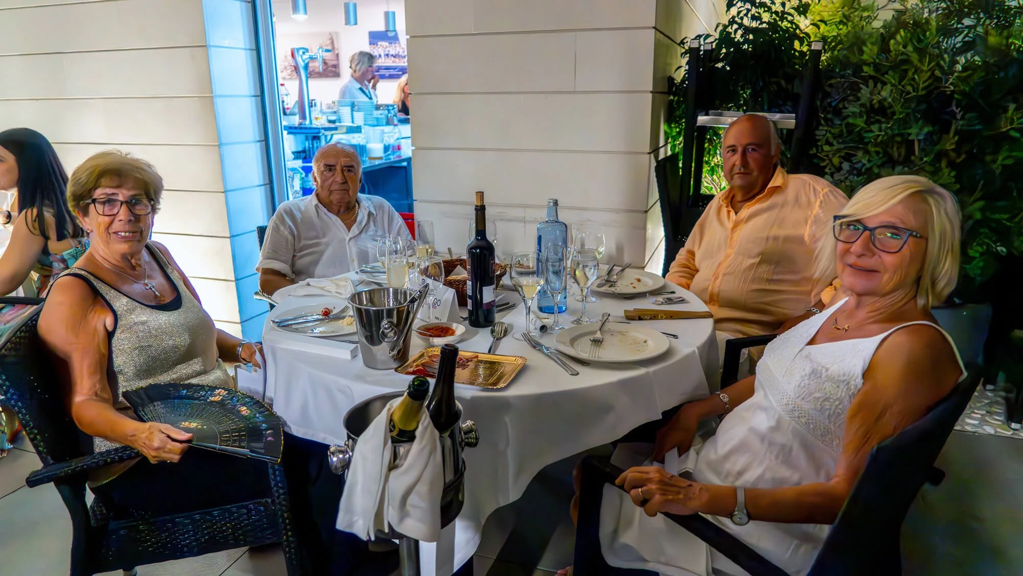 Grupo de cinco personas, sentadas alrededor de una mesa en un restaurante, con comida y bebidas en la mesa, algunos sonrientes, en un entorno con plantas y ventanas grandes.