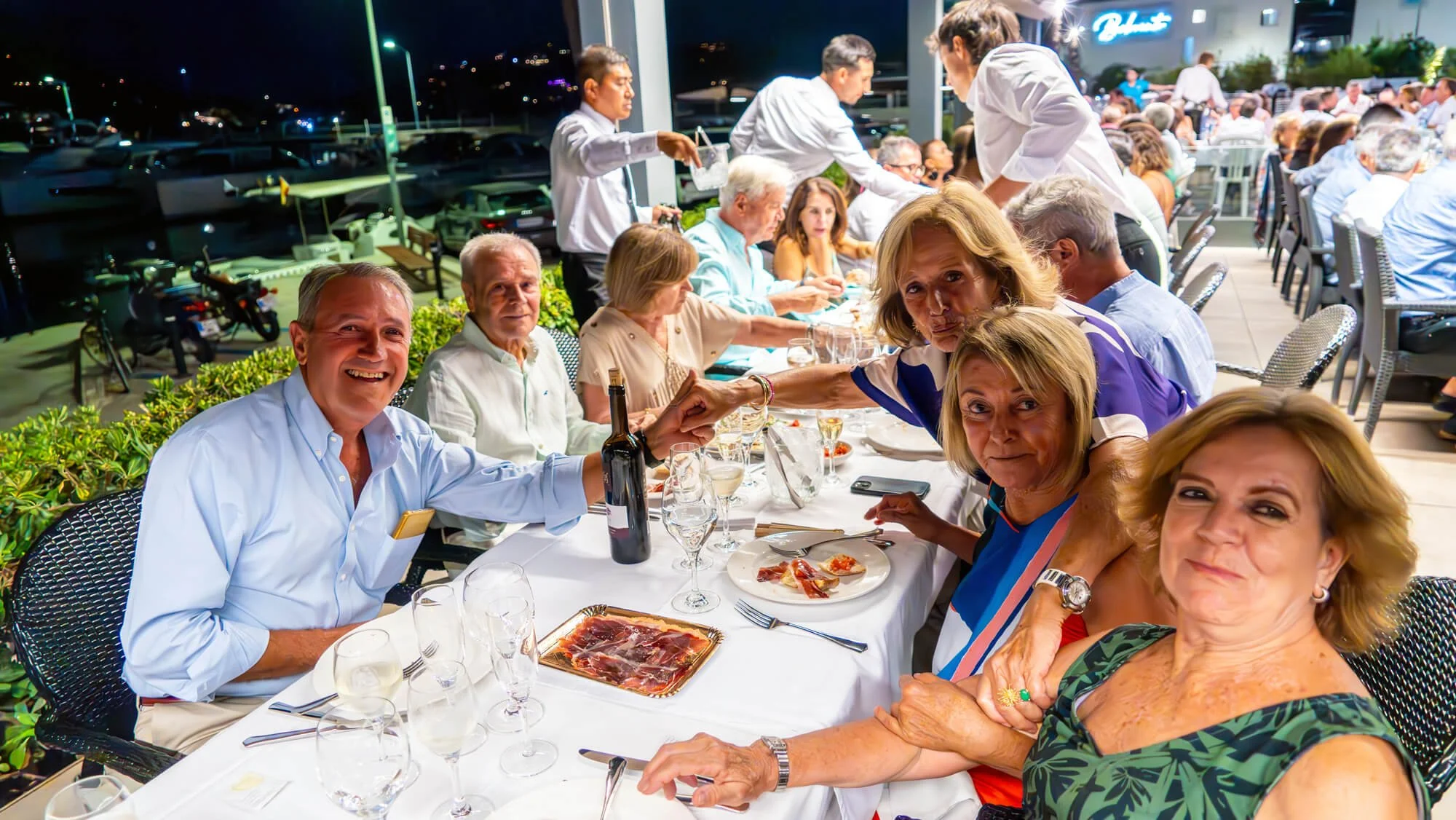 Grupo de personas celebrando una comida al aire libre en la noche, en una terraza con muchos comensales y un ambiente festivo.