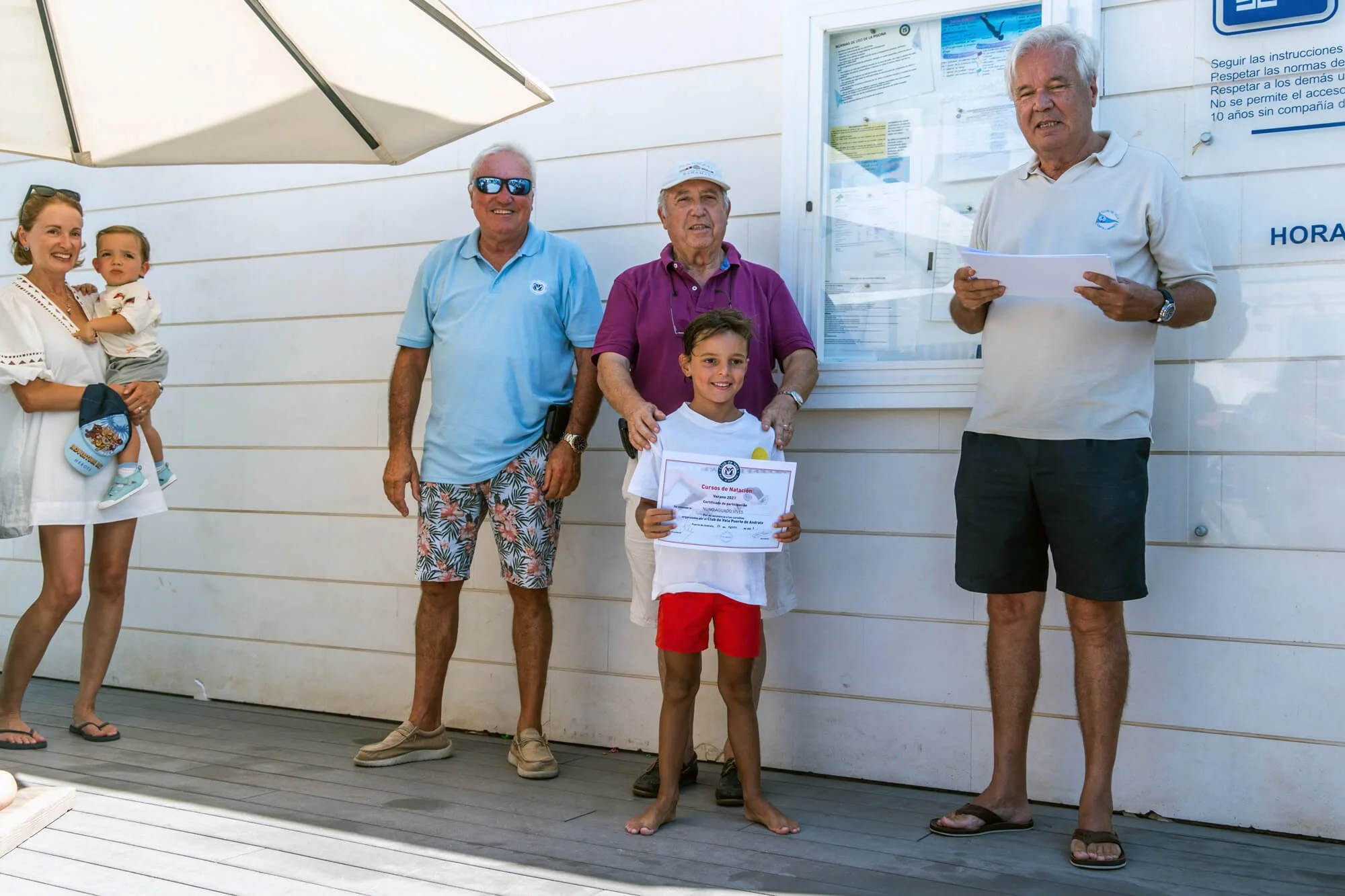 Grupo de personas en una ceremonia de premios, con un niño recibiendo diploma y adultos a su alrededor, algunos con camisetas blancas con logotipos y usando gafas de sol, en un espacio con pared blanca y cartel informativo.