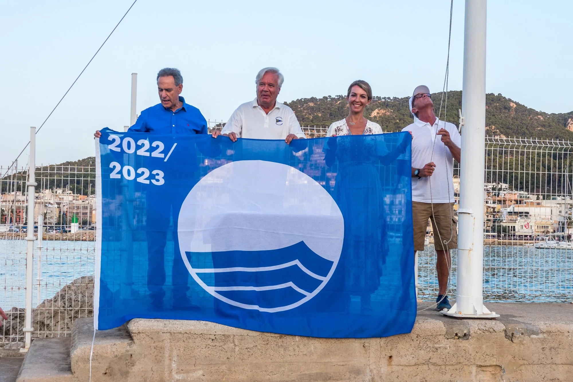 Cuatro personas sosteniendo una bandera azul con un símbolo de olas, en un área cercada junto al mar, con colinas verdes al fondo y un cielo despejado.