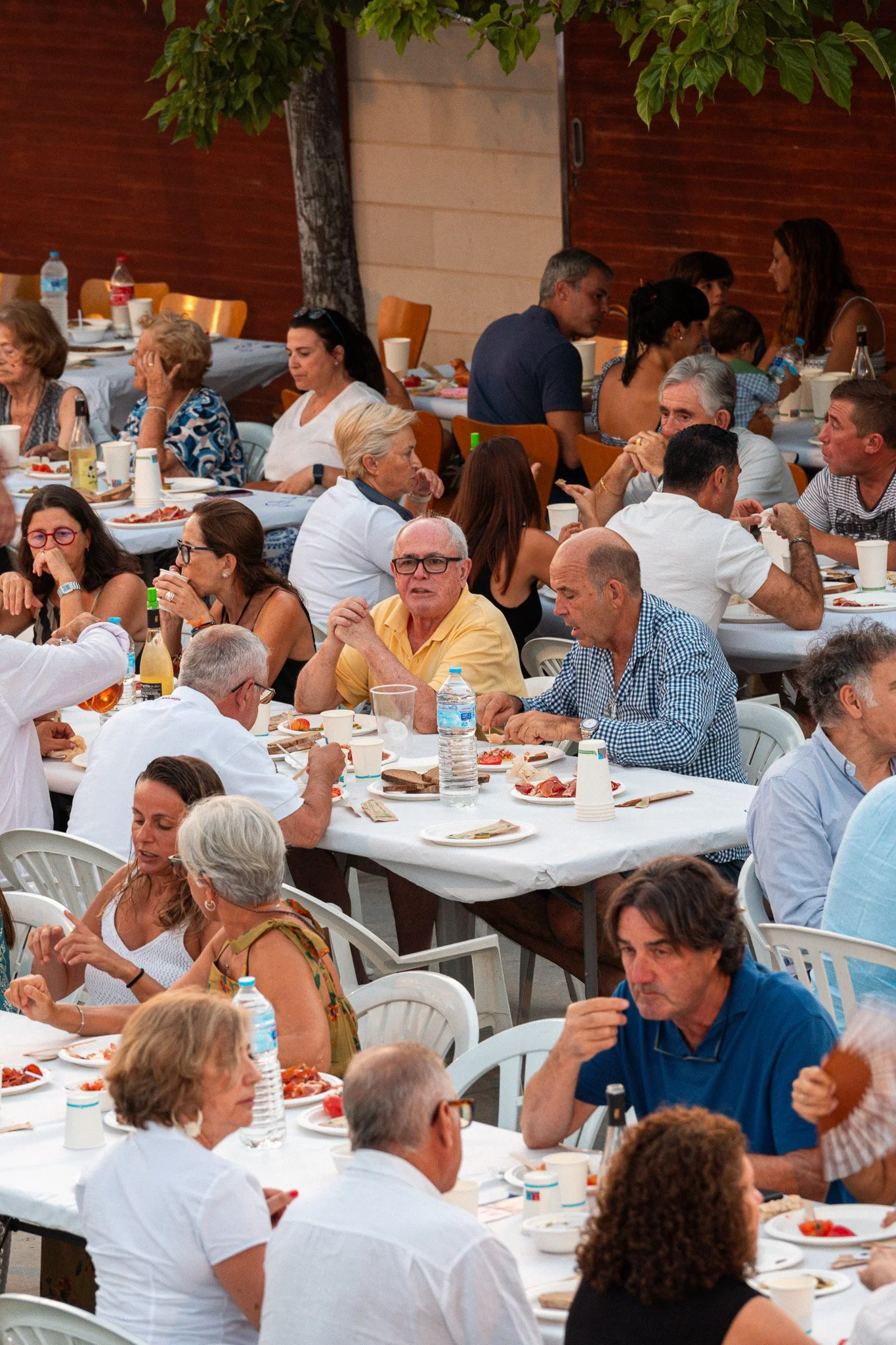 Personas reunidas en una comida o celebración al aire libre, sentadas en mesas con comida y bebidas, en un ambiente social y festivo.