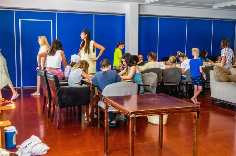 Niños y niñas en un área de juegos y actividades en un espacio cerrado, algunos sentados en mesas y otros de pie.