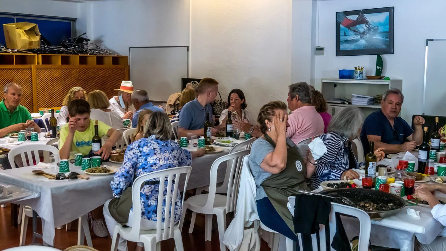 Personas comiendo en una cena familiar en un salón, mesas con platos, botellas de vino y vasos rojos y verdes, decoraciones y televisión en la pared