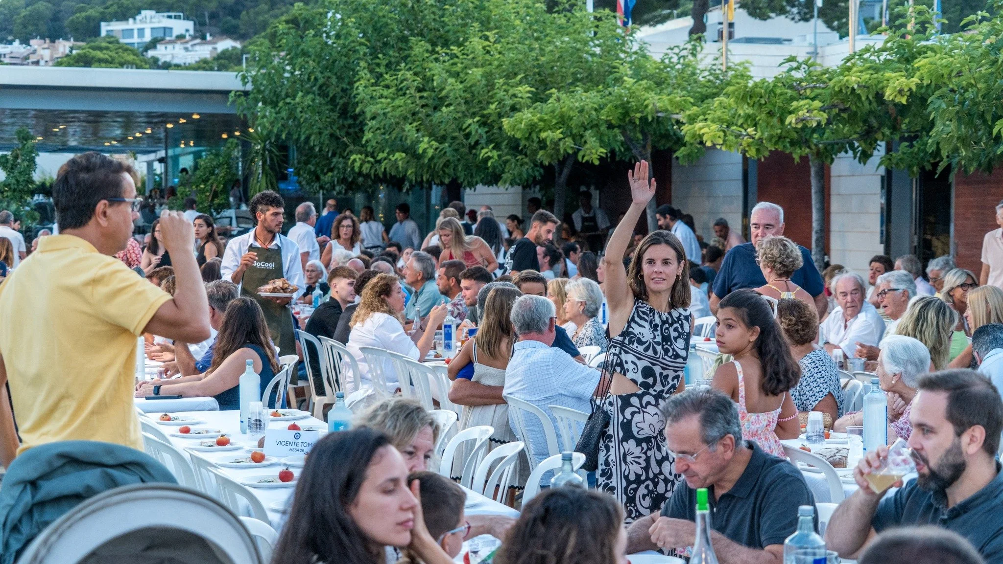 Una multitud de personas en una cena al aire libre, con muchas mesas y comida, en un ambiente animado y social, con árboles y edificios al fondo.
