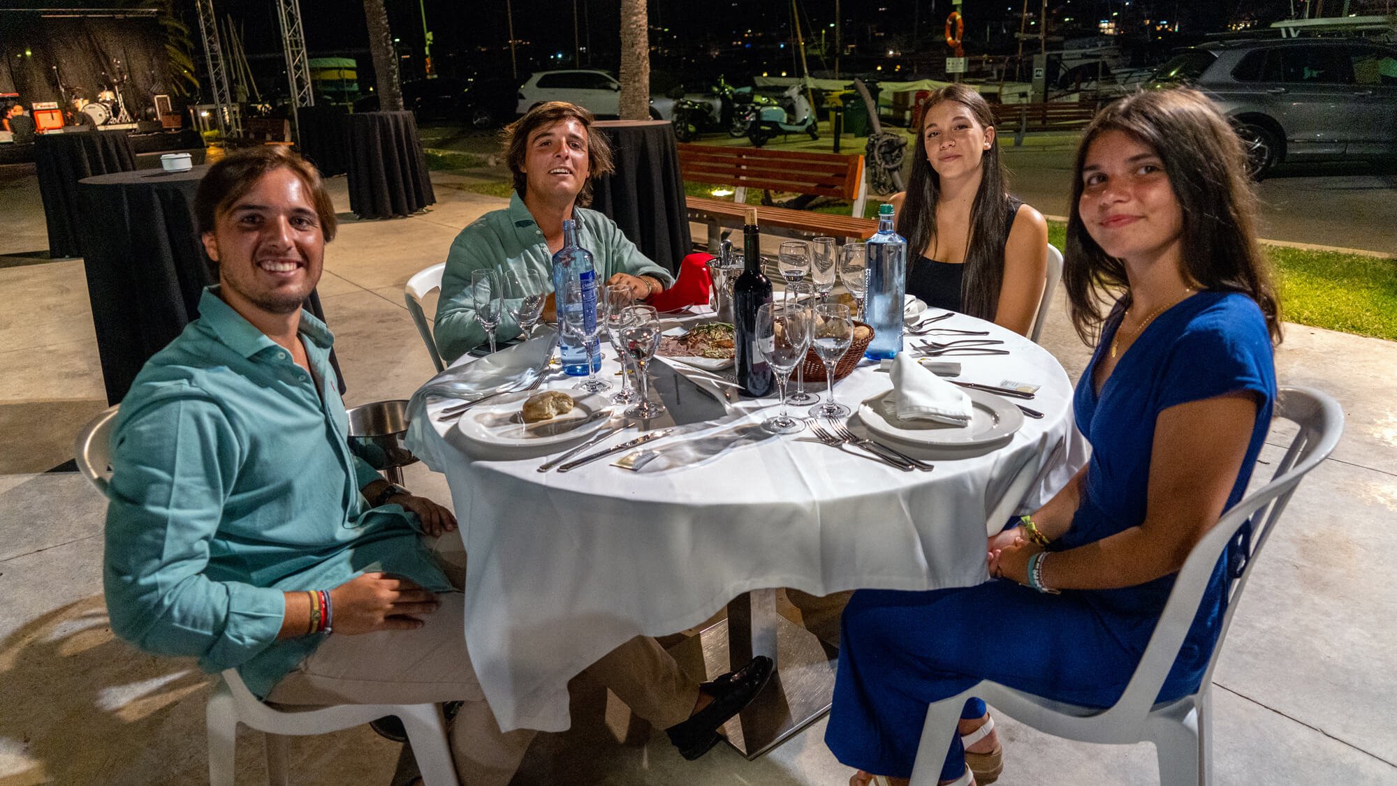 Cuatro personas jovenes sentadas en una mesa con comida en un restaurante al aire libre durante la noche, con detalles de la ciudad y autos en el fondo.