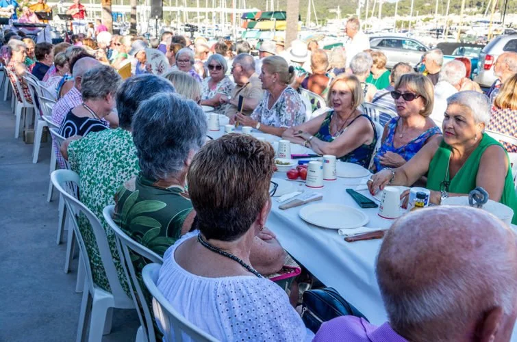 Grupo de personas mayores sentadas en una mesa larga al aire libre, participando en un evento social o comida comunitaria.