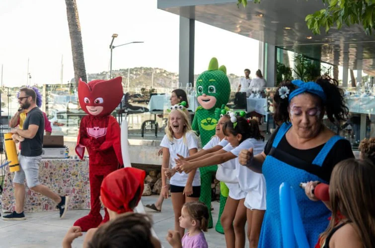 Niños y adultos disfrutando en un evento al aire libre con personajes de disfraces de superhéroes, junto a un muelle con barcos y una zona cubierta moderna.