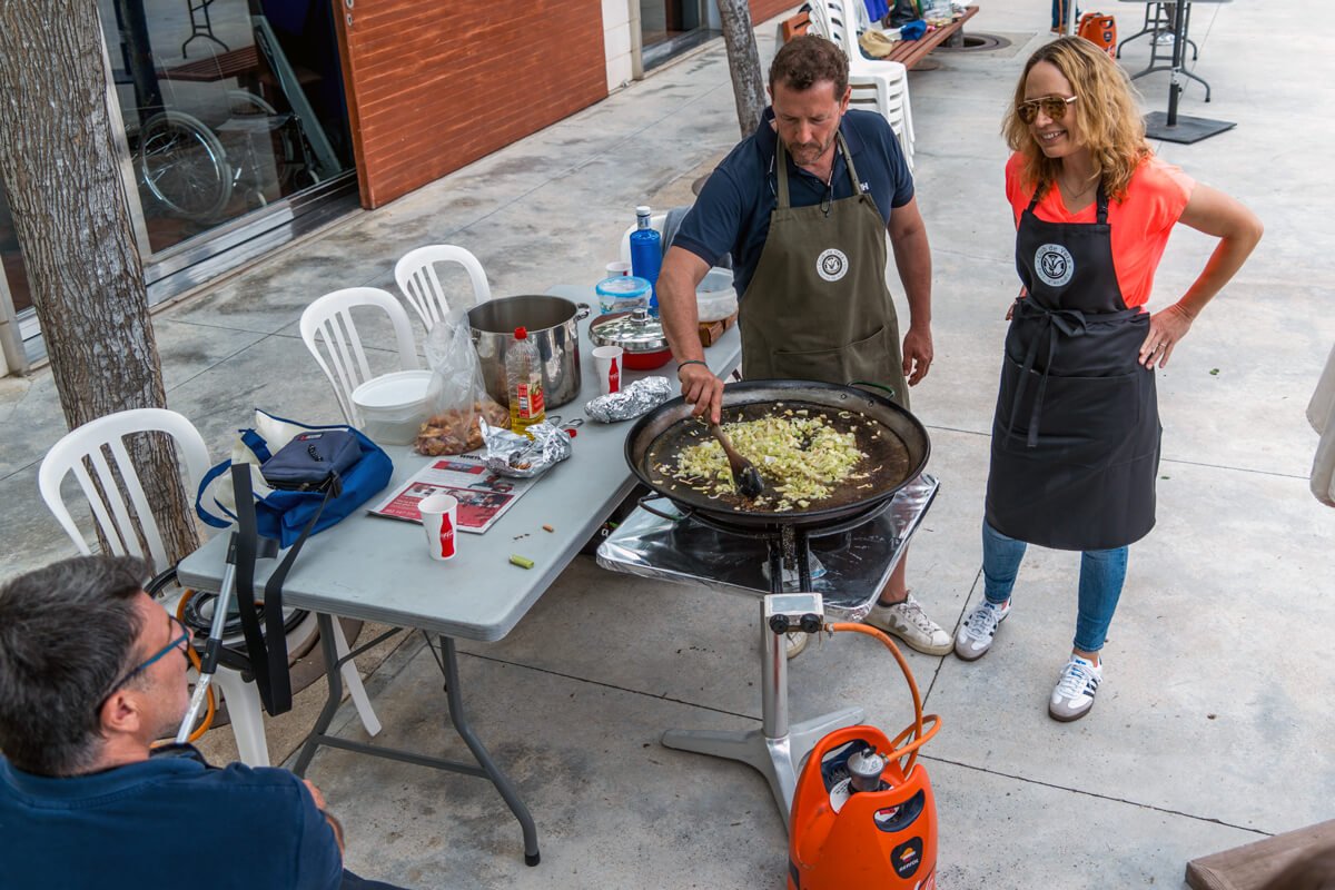 Dos personas cocinando en una parrilla en una mesa al aire libre mientras otro las observa, rodeadas de sillas y utensilios de cocina.
