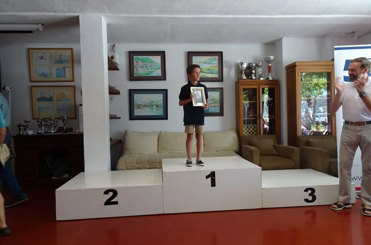 Un niño en el primer lugar del podio sostiene un trofeo, con un hombre a su derecha saludando y un fondo de cuadros y muebles en un salón.