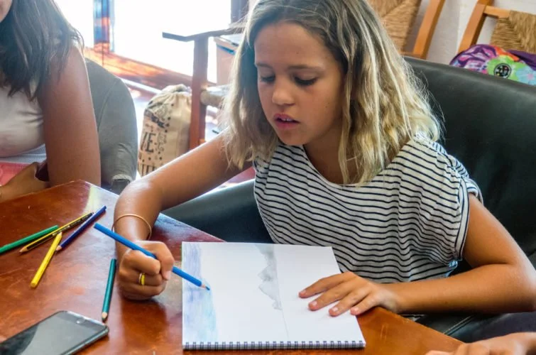 Una niña escribe o dibuja en un cuaderno con lápices de colores en una mesa de madera.