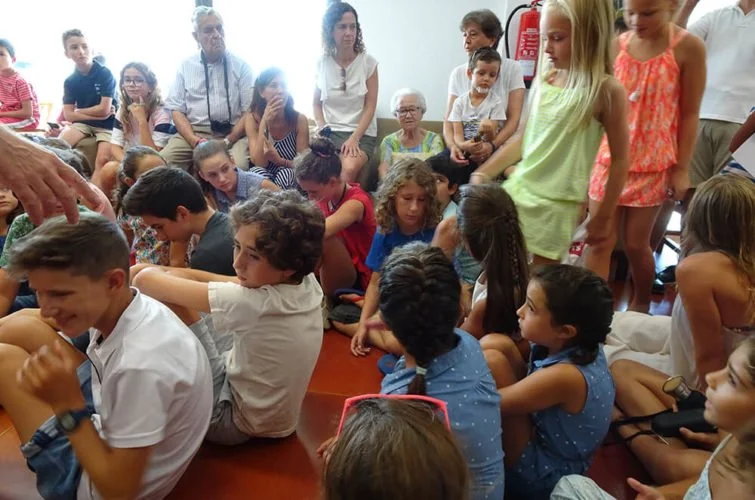 Grupo de niños y adultos sentados en un piso de madera en un ambiente interior, participando en una actividad grupal.