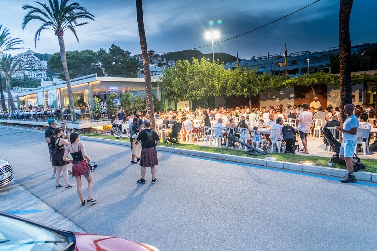 Cena al aire libre en la noche con muchas personas sentadas en mesas, palmeras y árboles, en un lugar con iluminación artificial, posible restaurante o cafetería.