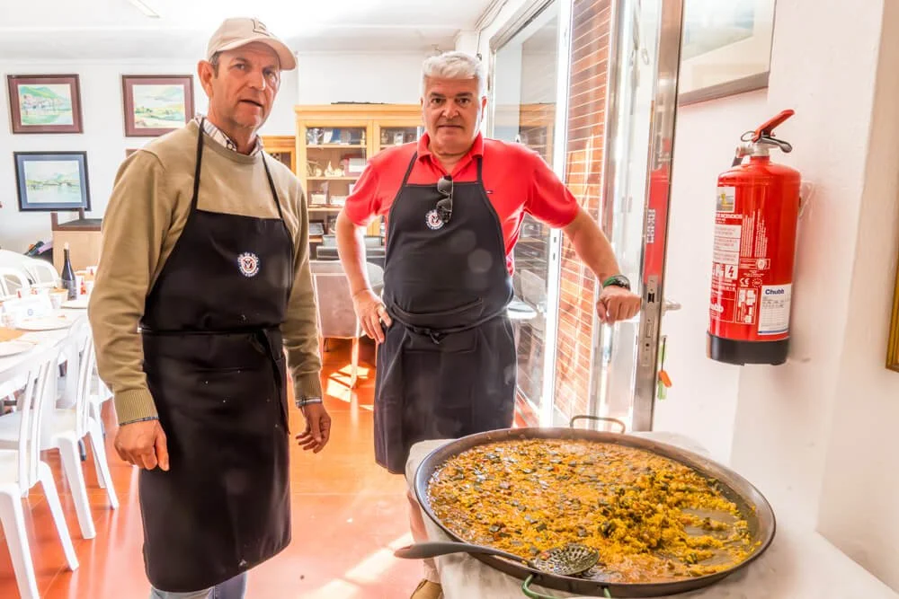 Dos hombres en una cocina, con delantales, junto a un plato grande de comida