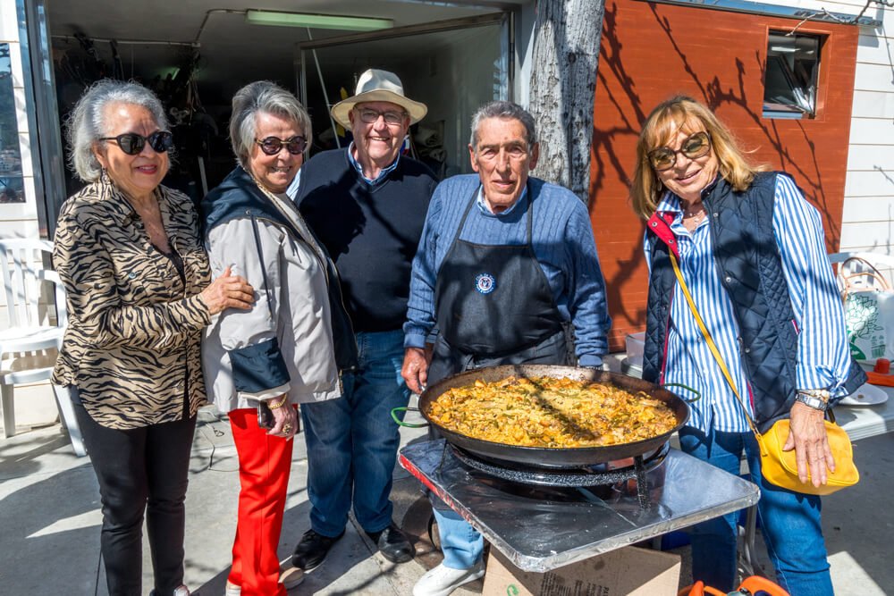 Cinco personas mayores posando con un plato grande de comida en una parrilla al aire libre.