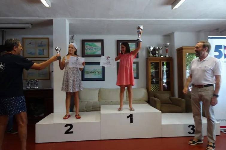 Dos niñas en el podio, recibiendo trofeos y diplomas en una ceremonia de premiación, con un adulto a la derecha y otro a la izquierda