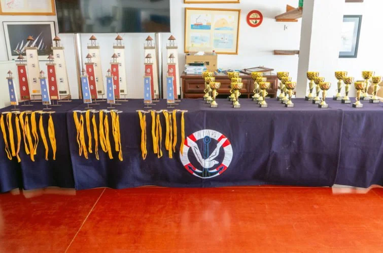 Mesas con trofeos y medallas en un evento, decoradas con un mantel azul y un logotipo del ancla en el centro.