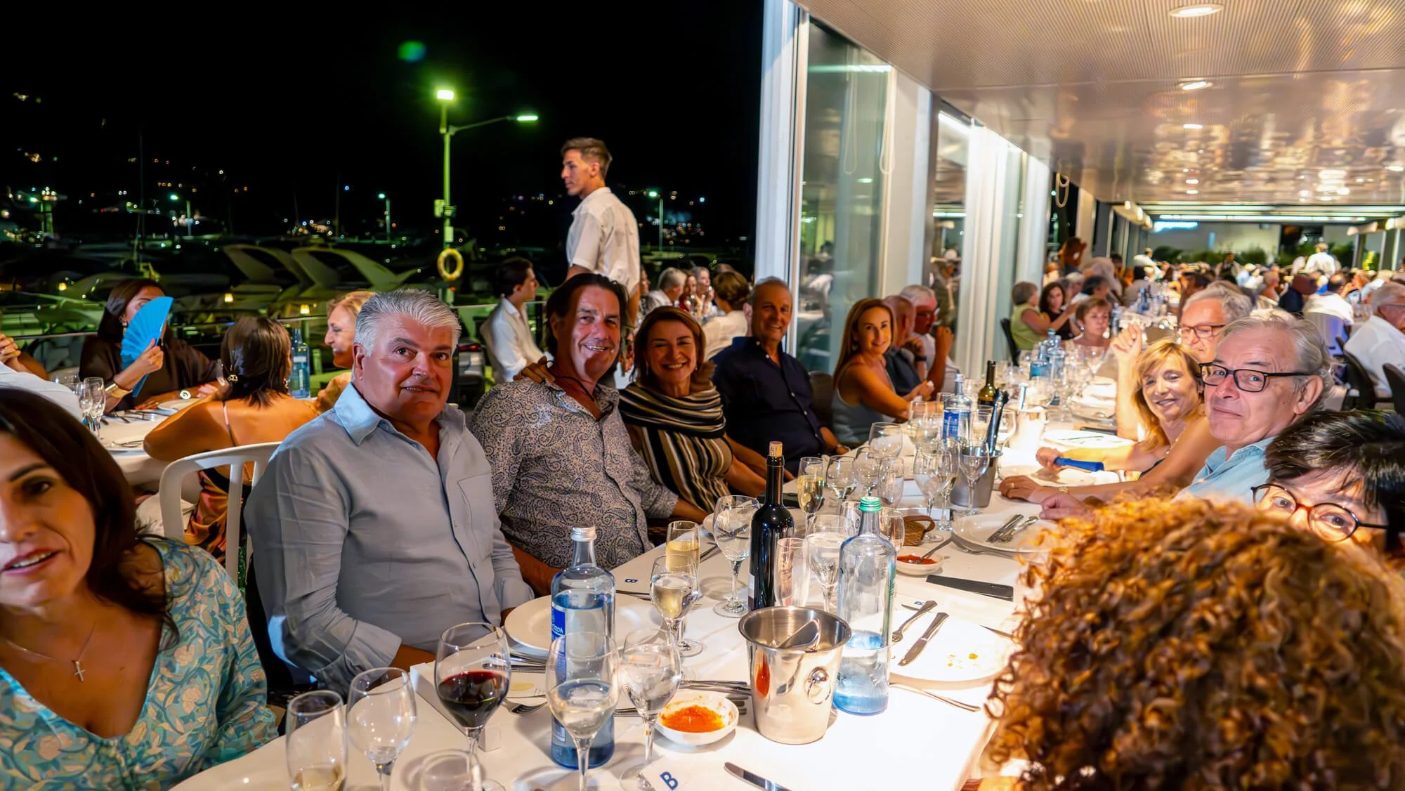 Cena de una cena en un restaurante con muchas personas, mucha comida y bebida sobre la mesa y ventanas que muestran un ambiente nocturno exterior.